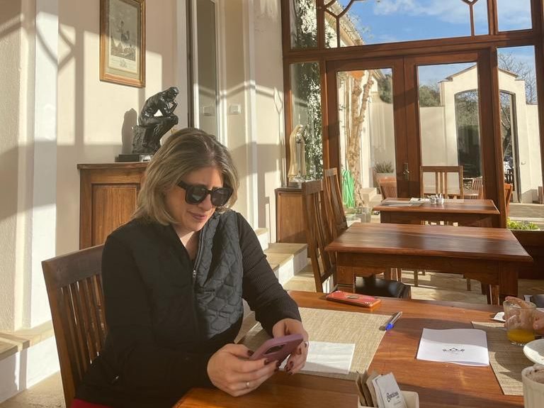 Woman with sunglasses sits in sunny room on her phone at a wooden table with big windows and sculpture in the background 